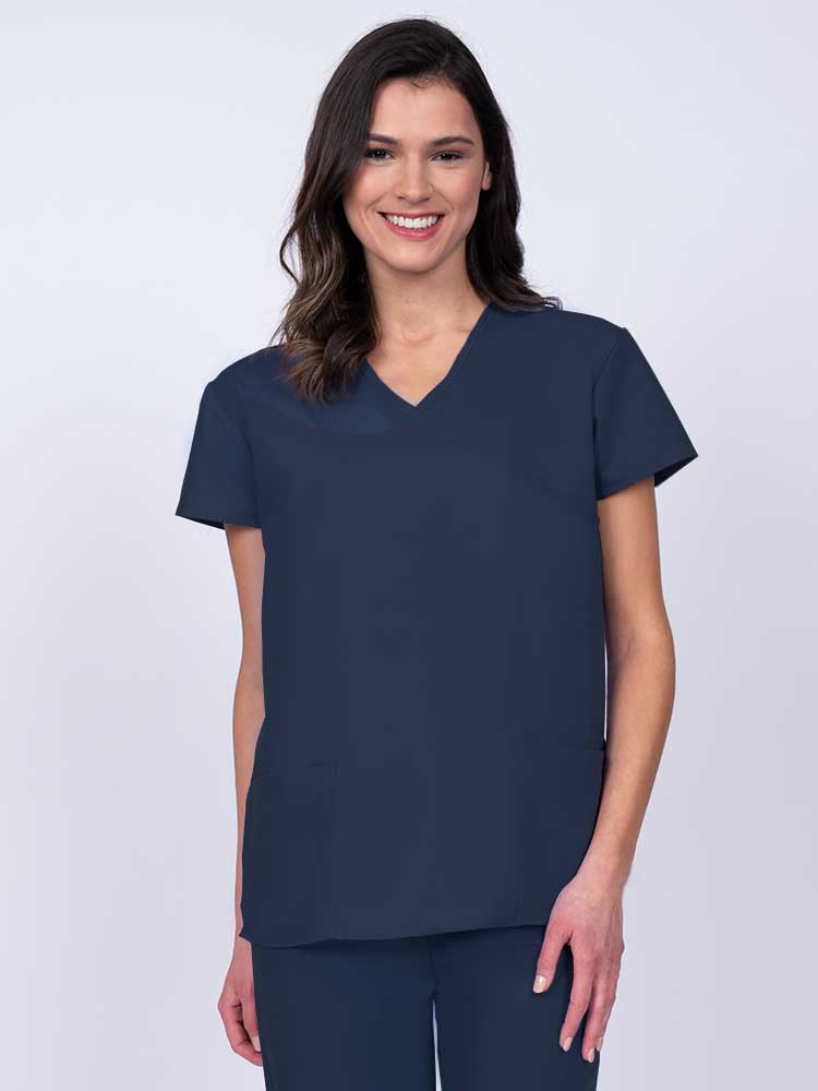 Young woman wearing a Luv Scrubs by MedWorks Women's Mock Wrap Scrub Top in navy featuring a Y-neckline and side slits for additional range of motion.