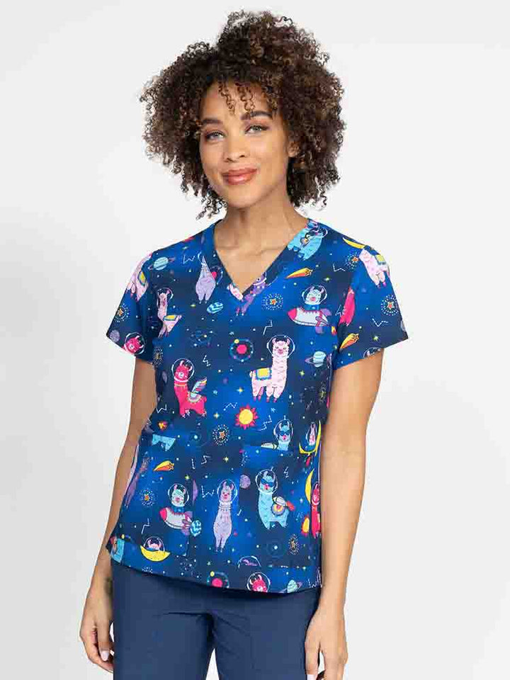 A young female Nurse Practitioner wearing a Meraki Sport Women's Print Scrub Top in "Space Llama" featuring a v-neckline & short sleeves.