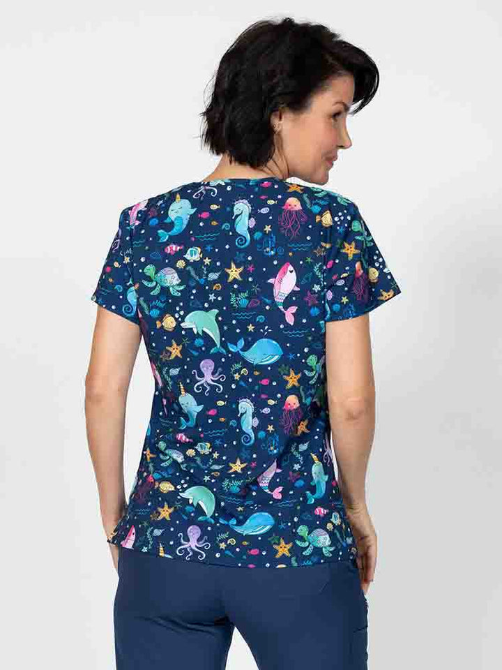 A young lady nurse wearing a Meraki Sport Women's Print Scrub Top in "Splish Splash" featuring shoulder yokes & side slits for additional range of motion.