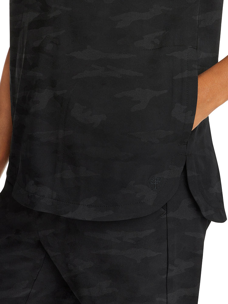 A young female Healthcare Professional wearing a Purple Label Women's Journey Camo Scrub Top in Black size Medium featuring side slits for additional mobility.