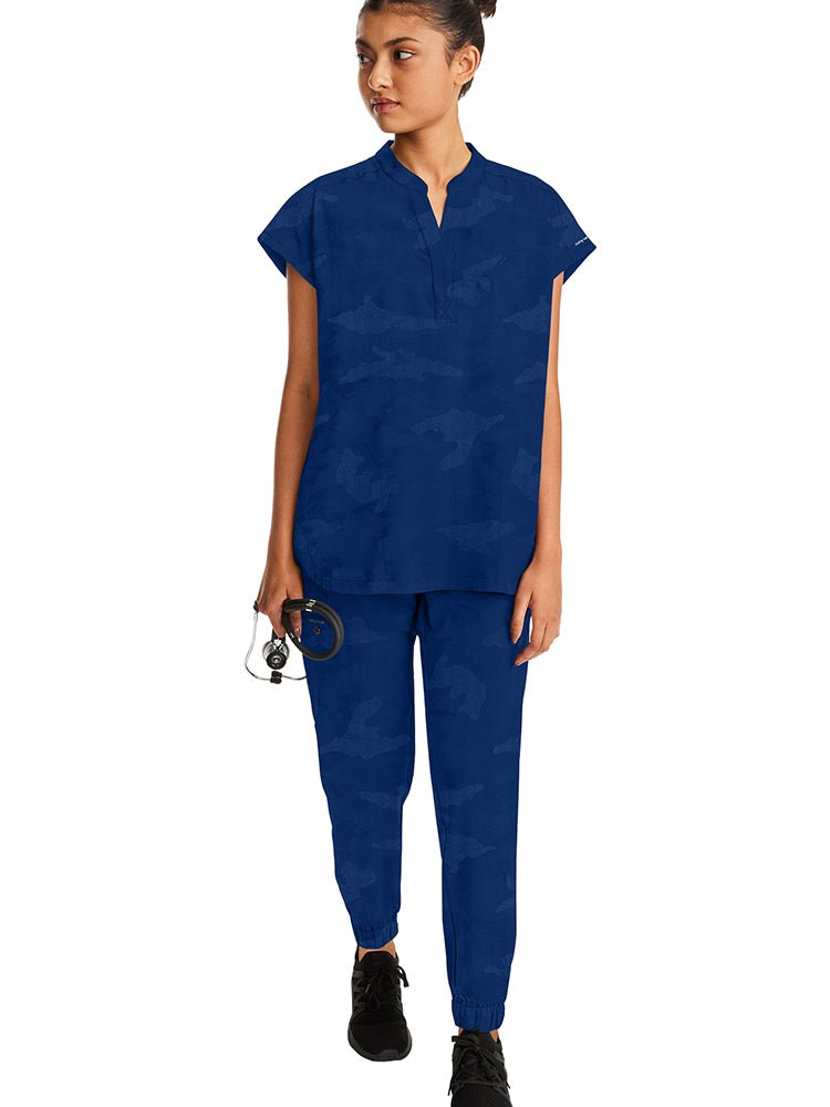 A female Nurse wearing a Purple Label Women's Journey Camo Scrub Top in Navy size XL featuring two front welt pockets.