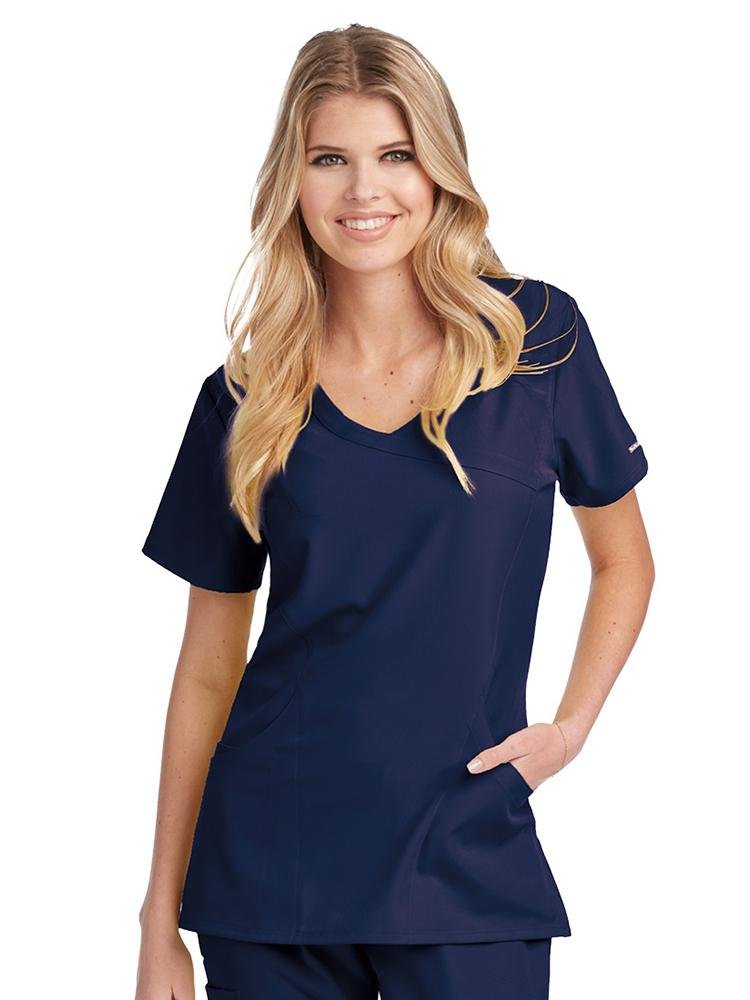 A female RN wearing a Skechers Women's Reliance Mock Wrap Scrub Top in Navy size small featuring two curved in-seam pockets & an interior mini pocket.
