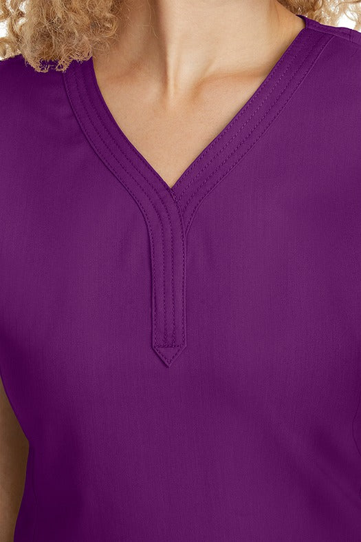 A young woman CNA wearing a Women's Jane V-Neck Scrub Top from Purple Label by Healing Hands in Eggplant featuring triple stitch detail at the neckline.