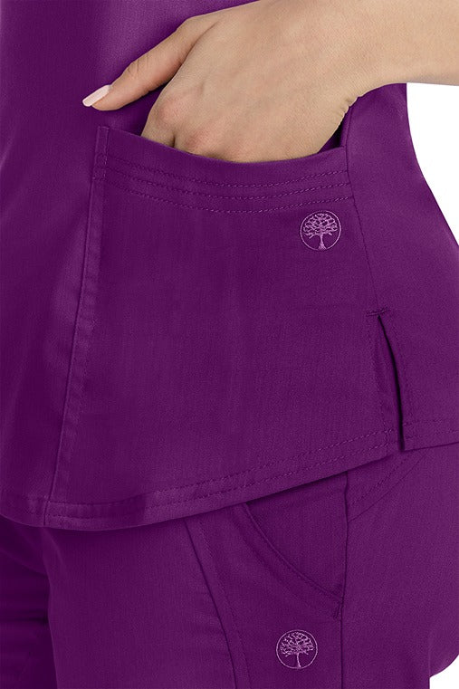 A female healthcare worker wearing a Purple Label Women's Jane V-Neck Scrub Top in Eggplant featuring triple needle stitch detail at the pockets.