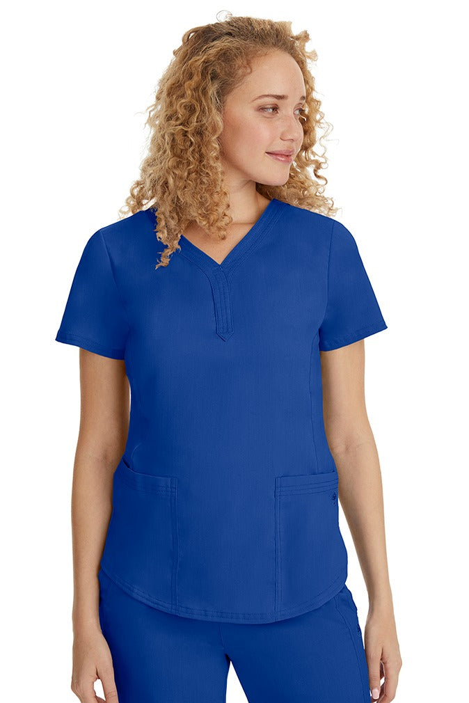 A young female healthcare professional wearing a Purple Label Women's Jane V-Neck Scrub Top in Galaxy Blue featuring 2 front patch pockets.