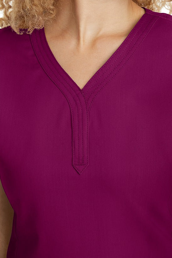 A young woman CNA wearing a Women's Jane V-Neck Scrub Top from Purple Label by Healing Hands in Wine featuring triple stitch detail at the neckline.