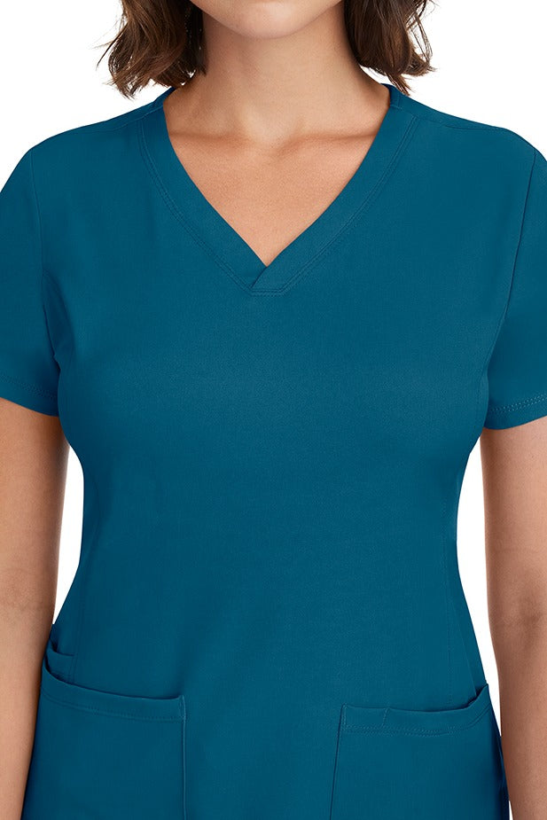 A young woman CNA wearing a HH-Works Women's Monica Multi-Pocket Scrub Top in Caribbean featuring front princess seaming to ensure a flattering fit.