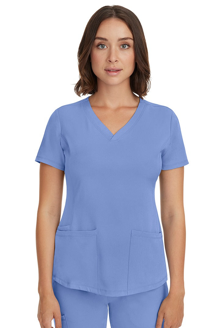 A young female LPN wearing a HH-Works Women's Monica Multi-Pocket Scrub Top in Ceil featuring short sleeves & a v-neckline.