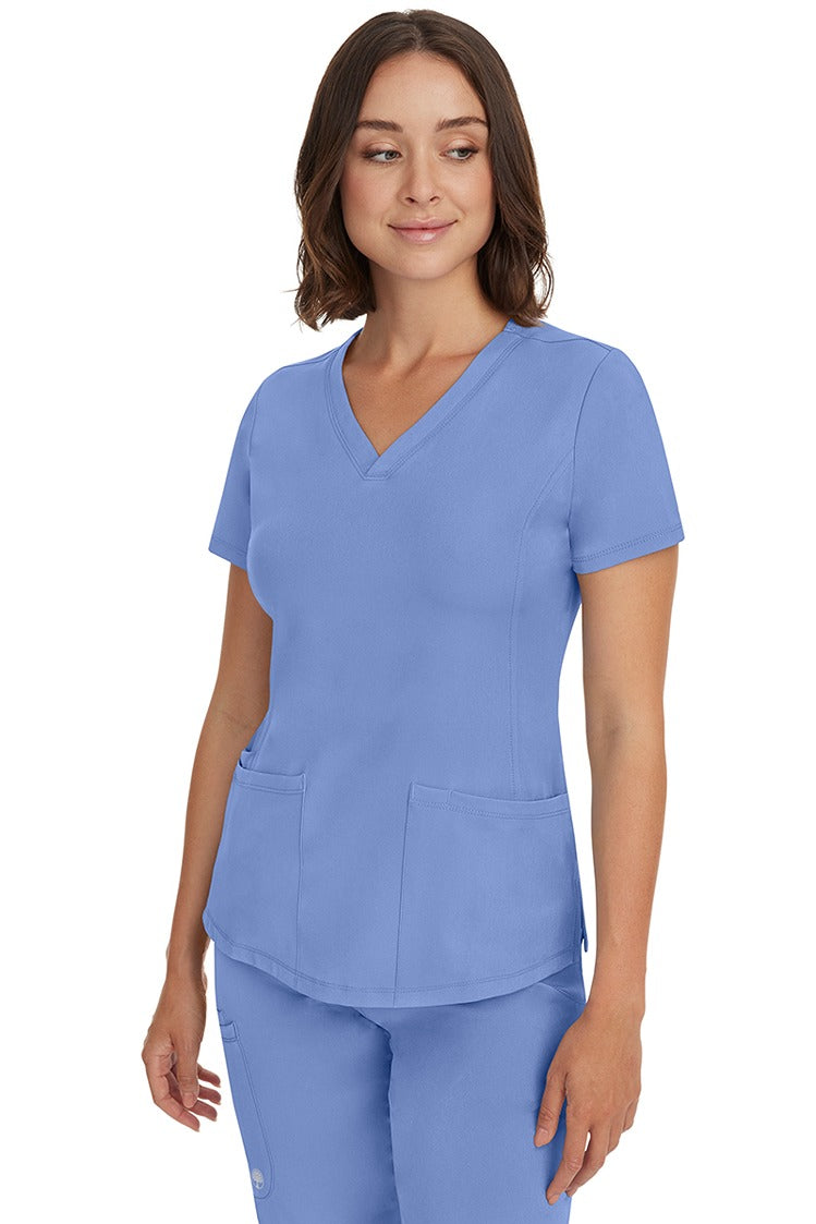 A young female healthcare professional wearing a HH-Works Women's Monica Multi-Pocket Scrub Top in Ceil featuring a center back length of 24".