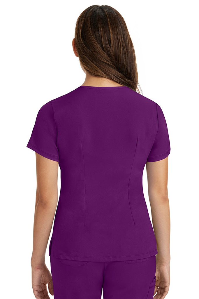 A female registered nurse wearing a Women's Monica Multi-Pocket Scrub Top from HH Works in Eggplant featuring back princess darts for shaping.