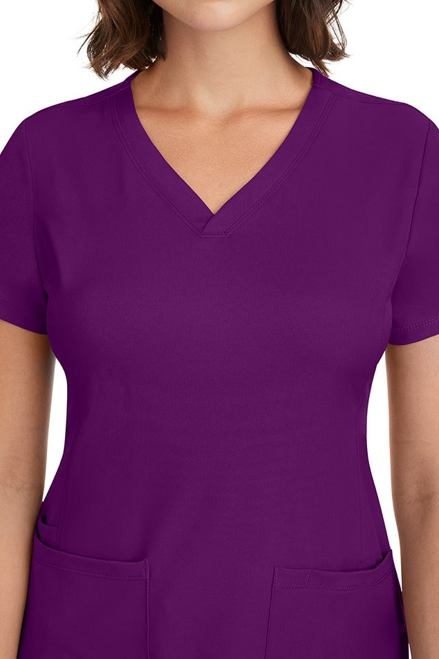A young woman CNA wearing a HH-Works Women's Monica Multi-Pocket Scrub Top in Eggplant featuring front princess seaming to ensure a flattering fit.