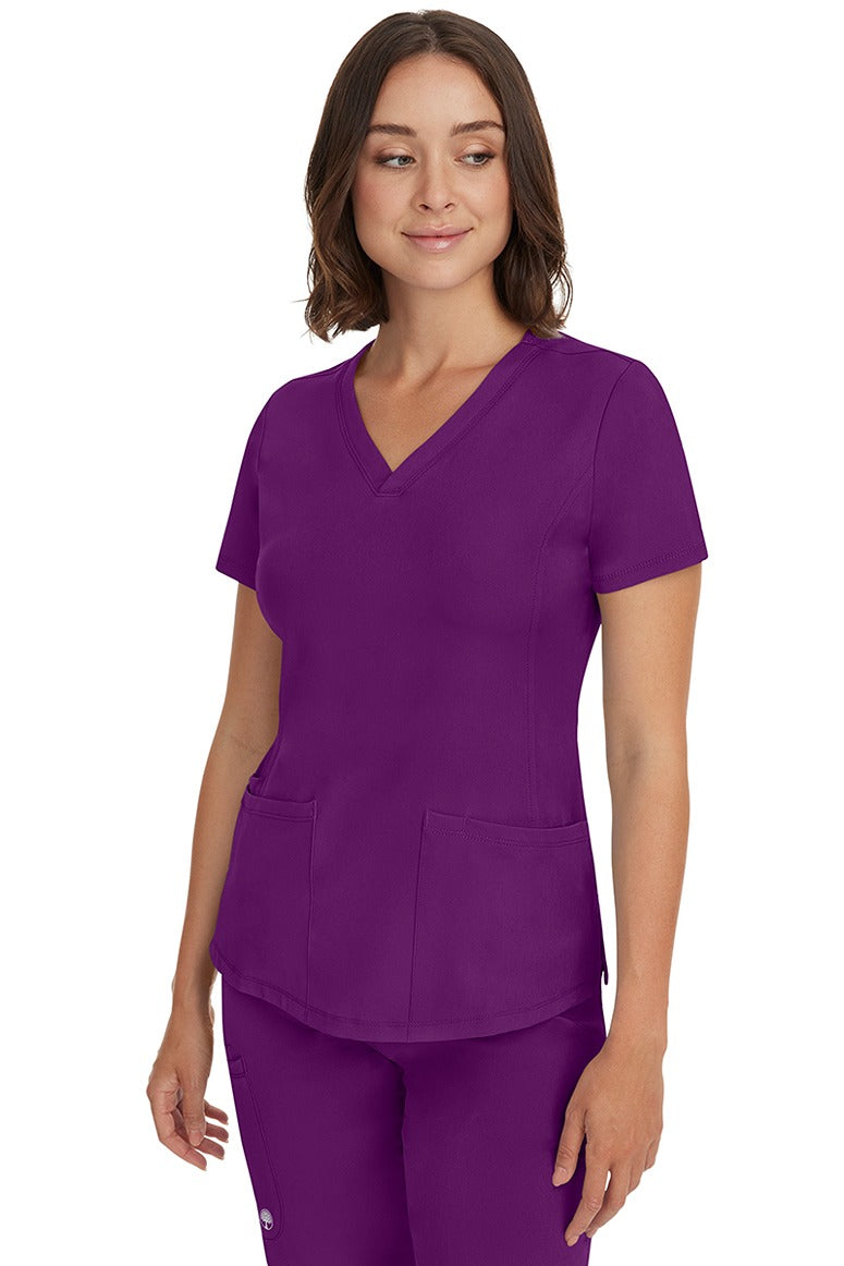A young female healthcare professional wearing a HH-Works Women's Monica Multi-Pocket Scrub Top Eggplant  featuring a center back length of 24".