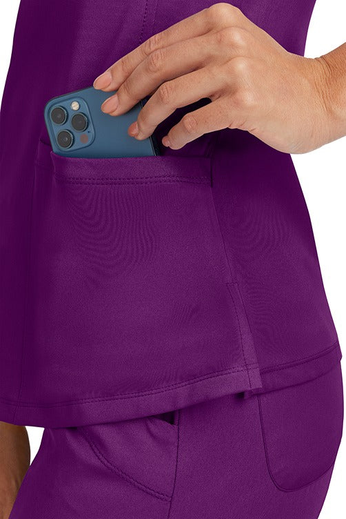 A young Home Care Registered Nurse wearing a HH-Works Women's Monica Multi-Pocket Scrub Top in Eggplant featuring side slits for additional range of motion.