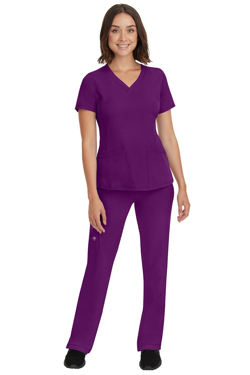 A young female nurse wearing a Women's Monica Multi-Pocket Scrub Top from HH Works in Eggplant featuring unique stretch fabric made of 91% polyester & 9% spandex.