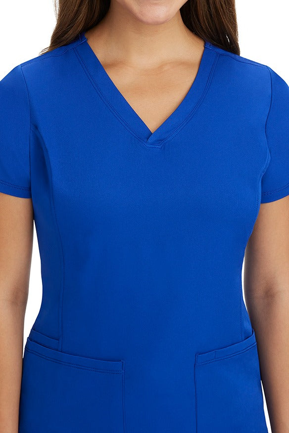 A young woman CNA wearing a HH-Works Women's Monica Multi-Pocket Scrub Top in Galaxy Blue  featuring front princess seaming to ensure a flattering fit.
