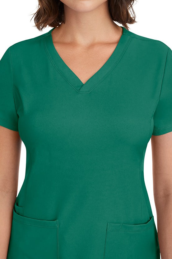 A young woman CNA wearing a HH-Works Women's Monica Multi-Pocket Scrub Top in Hunter Green featuring front princess seaming to ensure a flattering fit.
