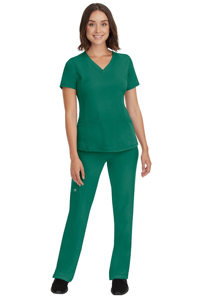 A young female nurse wearing a Women's Monica Multi-Pocket Scrub Top from HH Works in Hunter Green featuring unique stretch fabric made of 91% polyester & 9% spandex.