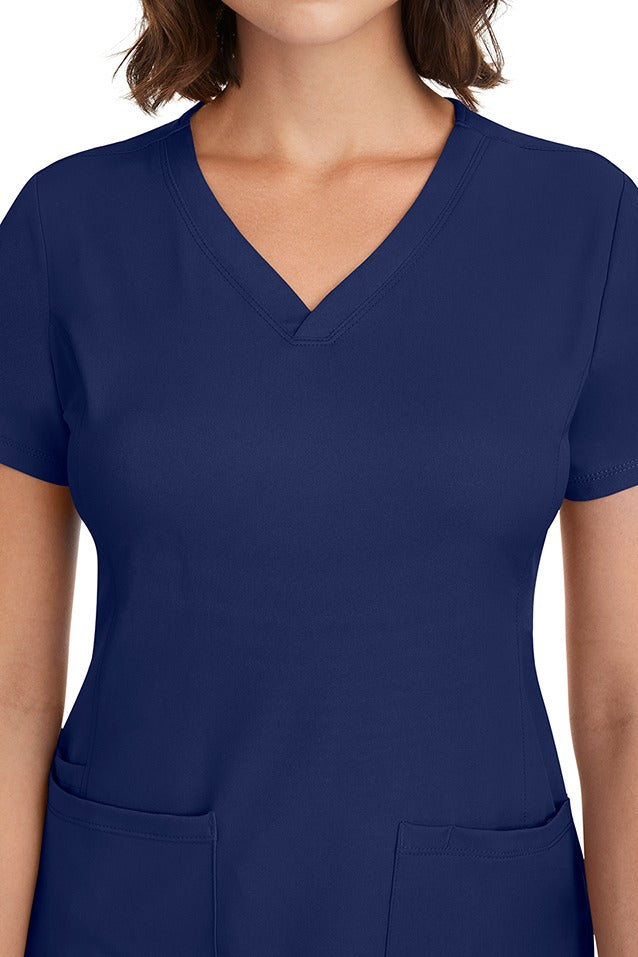 A young woman CNA wearing a HH-Works Women's Monica Multi-Pocket Scrub Top in Navy featuring front princess seaming to ensure a flattering fit.