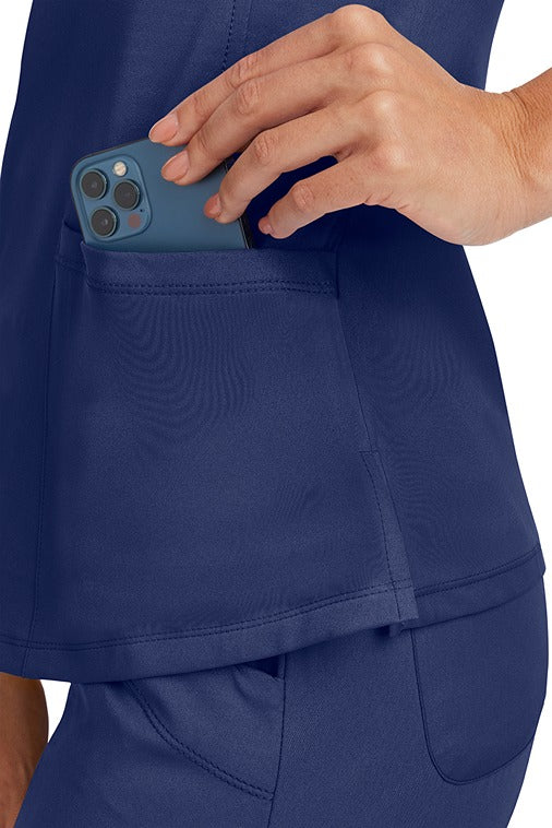A young Home Care Registered Nurse wearing a HH-Works Women's Monica Multi-Pocket Scrub Top in Navy featuring side slits for additional range of motion.