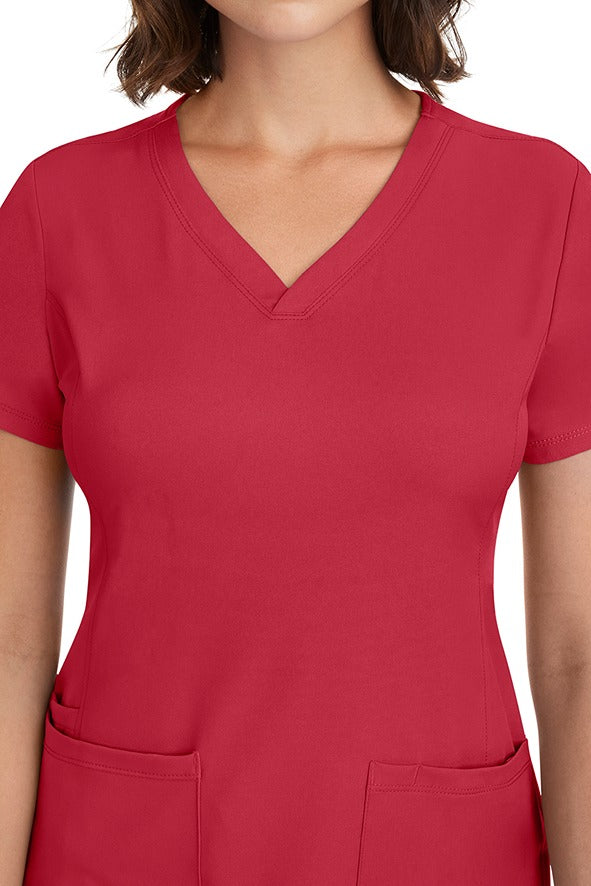 A young woman CNA wearing a HH-Works Women's Monica Multi-Pocket Scrub Top in Red featuring front princess seaming to ensure a flattering fit.