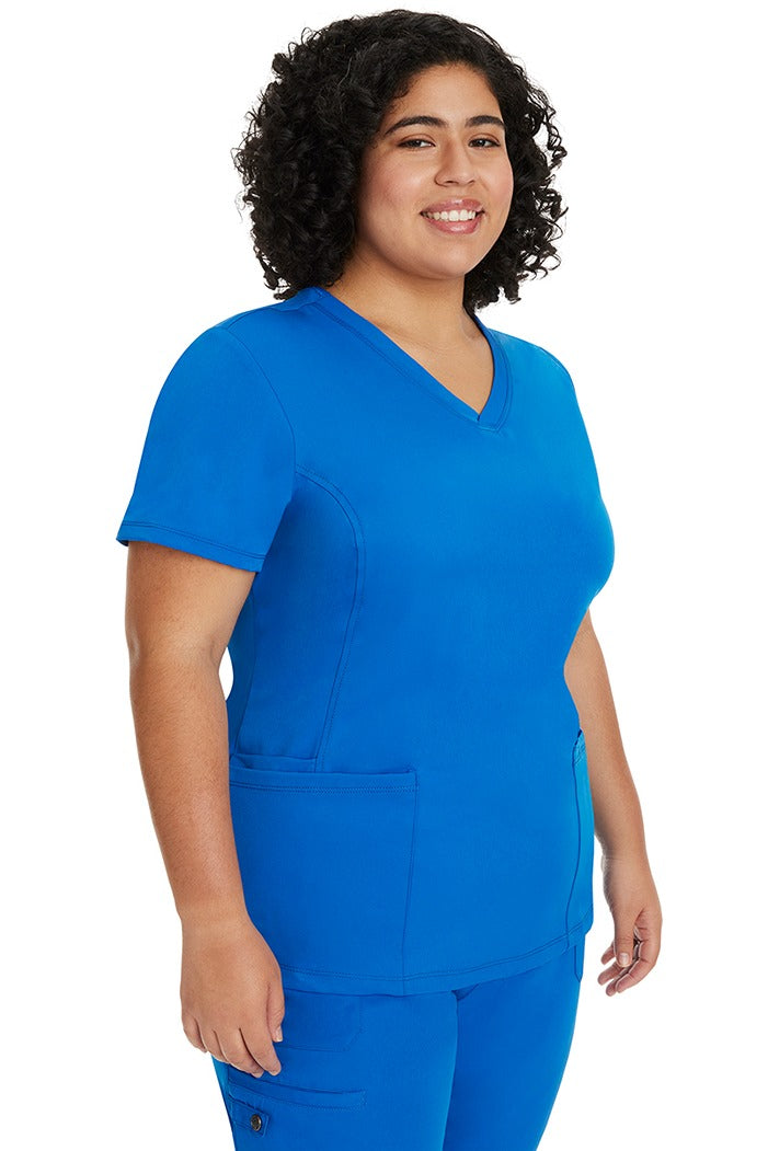 A young female healthcare professional wearing a HH-Works Women's Monica Multi-Pocket Scrub Top in Royal featuring a center back length of 24".