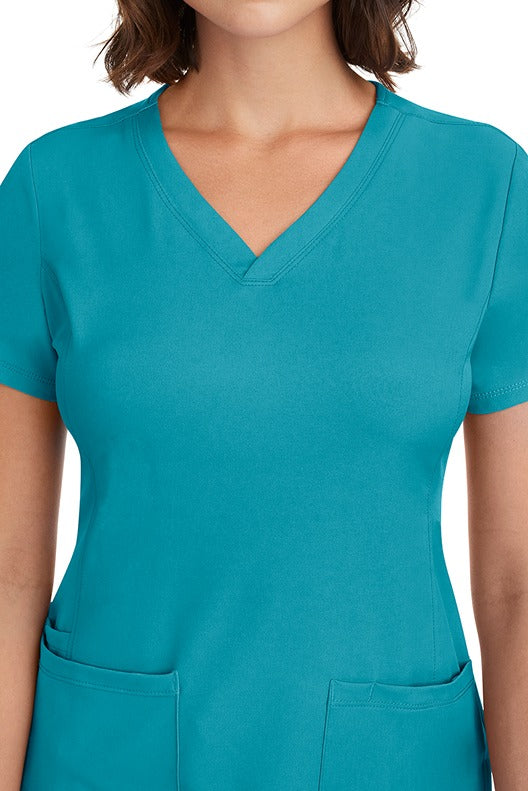 A young woman CNA wearing a HH-Works Women's Monica Multi-Pocket Scrub Top in Teal featuring front princess seaming to ensure a flattering fit.