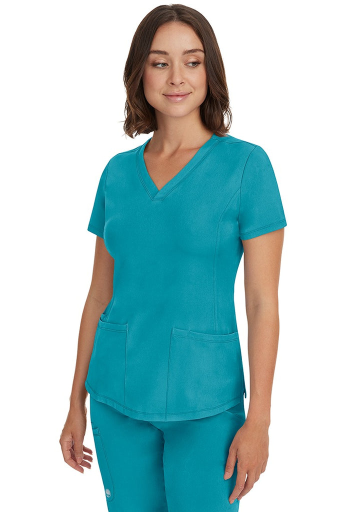A young female healthcare professional wearing a HH-Works Women's Monica Multi-Pocket Scrub Top in Teal featuring a center back length of 24".