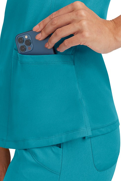 A young Home Care Registered Nurse wearing a HH-Works Women's Monica Multi-Pocket Scrub Top in Teal featuring side slits for additional range of motion.