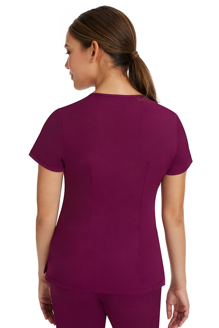 A female registered nurse wearing a Women's Monica Multi-Pocket Scrub Top from HH Works in Wine featuring back princess darts for shaping.