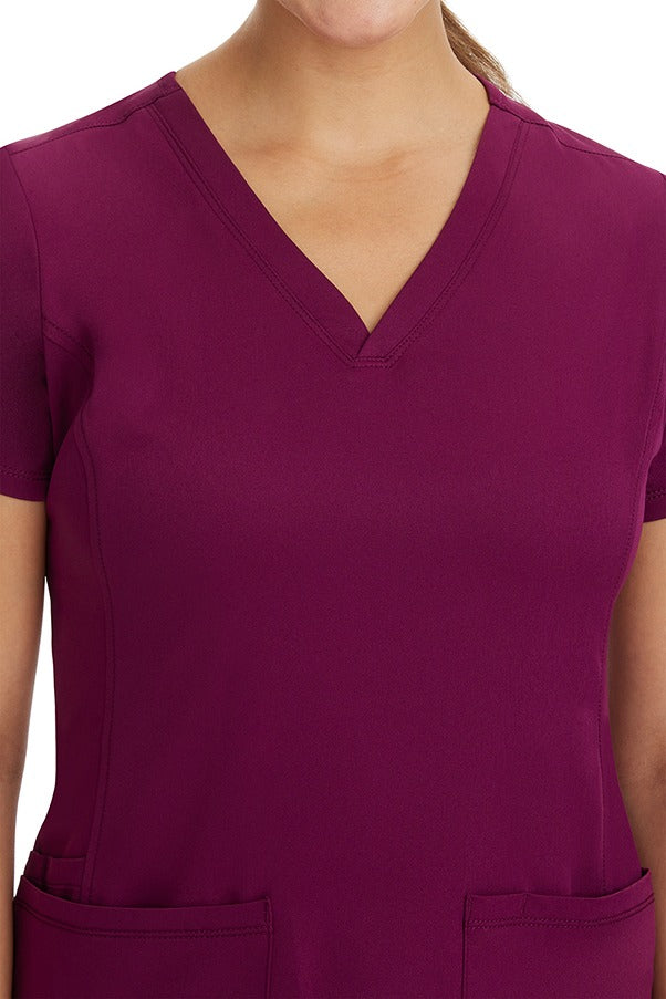 A young woman CNA wearing a HH-Works Women's Monica Multi-Pocket Scrub Top in Wine featuring front princess seaming to ensure a flattering fit.