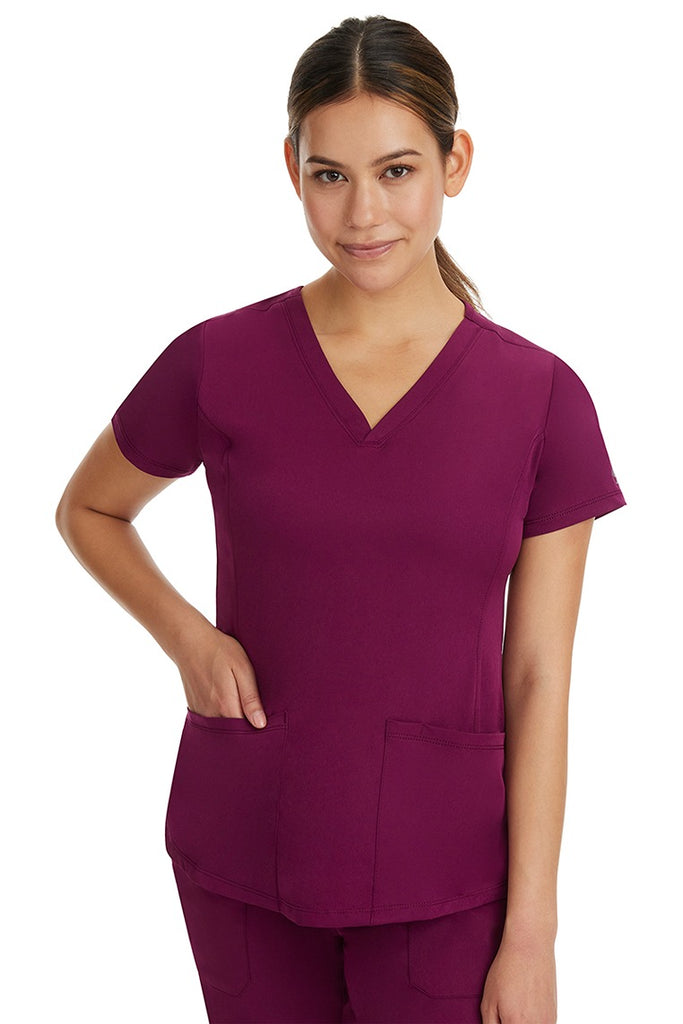 A young female healthcare professional wearing a HH-Works Women's Monica Multi-Pocket Scrub Top in Wine featuring a center back length of 24".