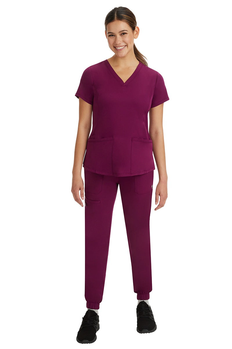 A young female Medical Assistant wearing a Women's Monica Multi-Pocket Scrub Top from HH Works in Wine featuring unique stretch fabric made of 91% polyester & 9% spandex.
