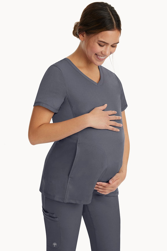 A young woman wearing an HH-Works Women's Mila Maternity V-Neck Scrub Top in Pewter featuring adjustable side panels.