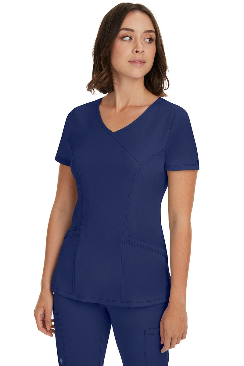 A young woman wearing a Women's Madison Mock Wrap Scrub Top from HH Works in Navy featuring a medium center back length of 24".