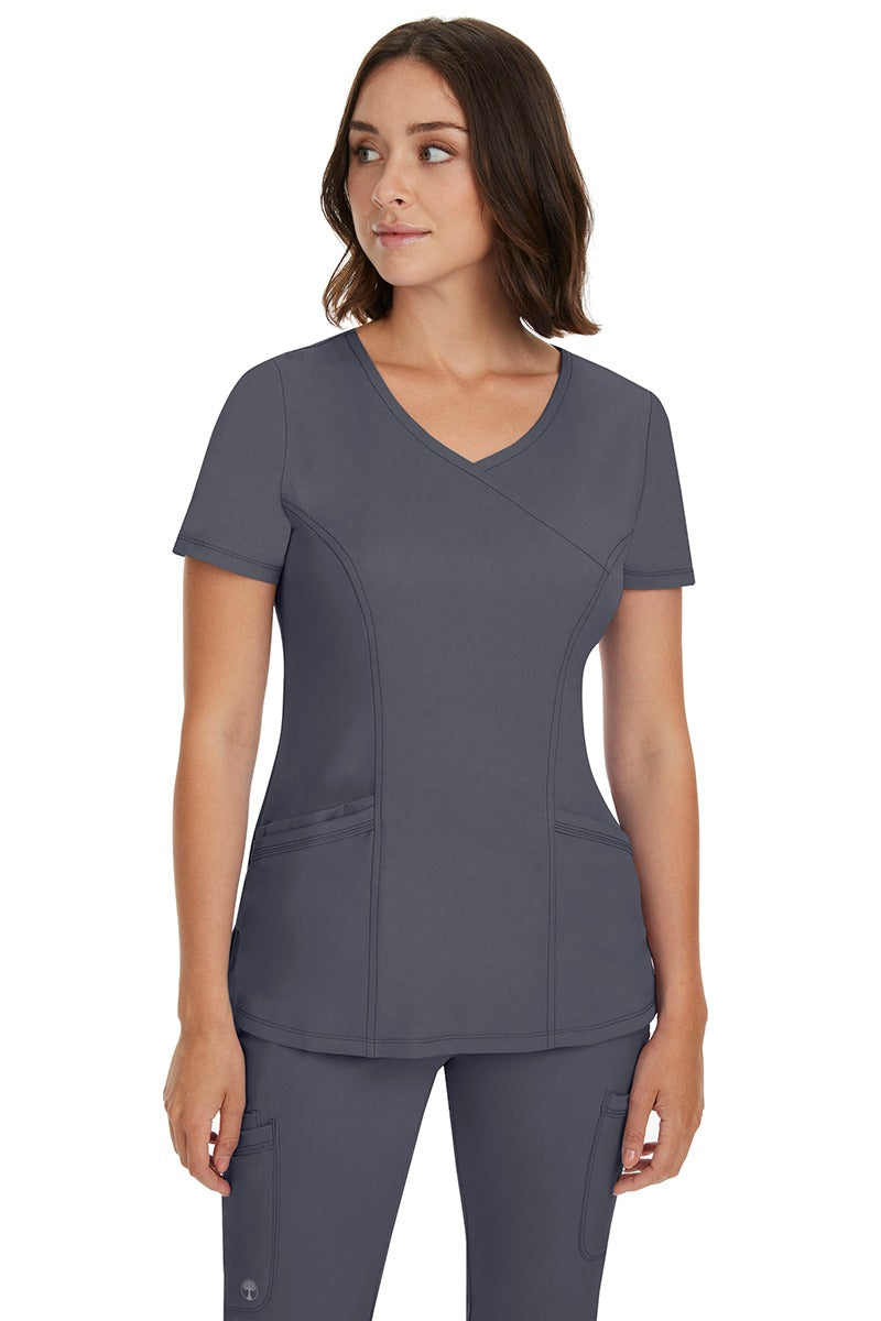A young Home Care Registered Nurse wearing a HH-Works Women's Madison Mock Wrap Scrub Top in Pewter featuring a total of 4 pockets for all of your on the go storage needs.
