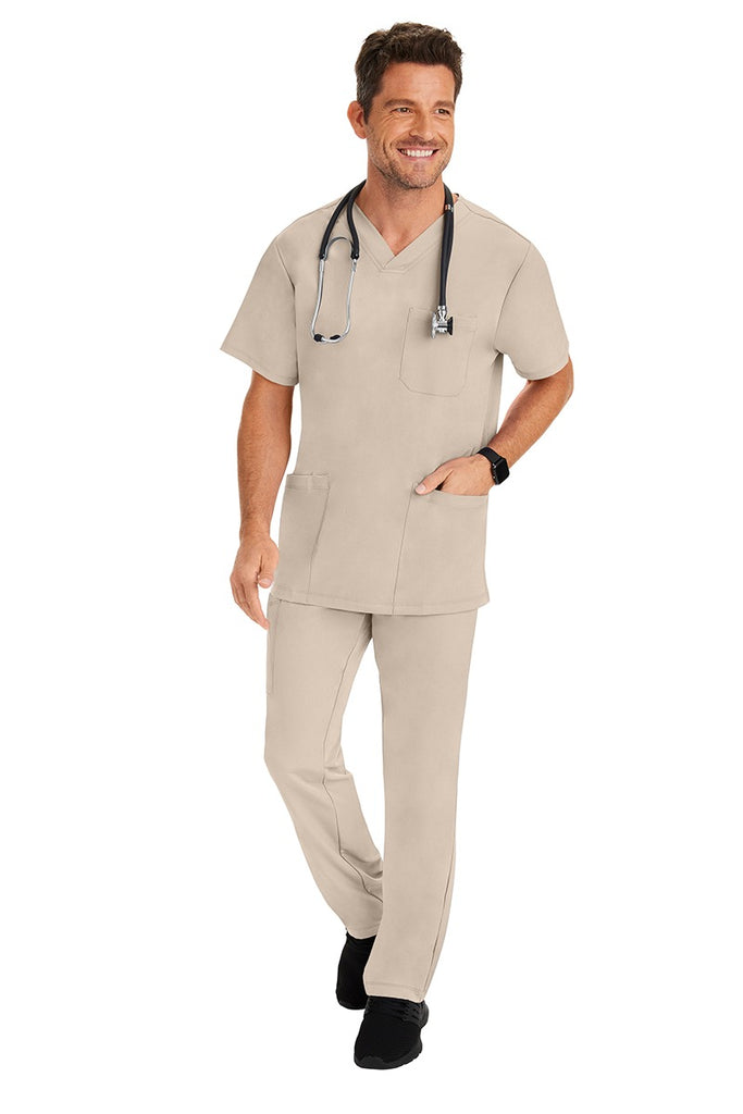 A young male Home Care Registered Nurse wearing an HH-Works Men's Matthew V-Neck Scrub Top in Khaki featuring a fade resistant fabric to ensure a long-lasting comfortable fit.