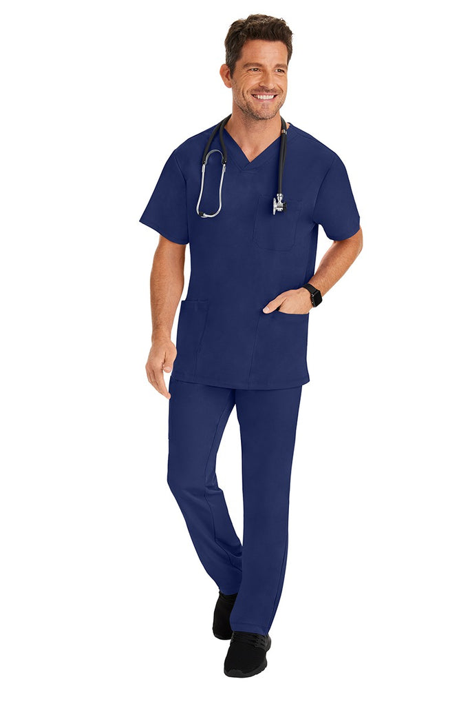 A young male Home Care Registered Nurse wearing an HH-Works Men's Matthew V-Neck Scrub Top in Navy featuring a fade resistant fabric to ensure a long-lasting comfortable fit.