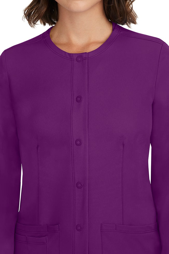 A female LPN wearing a Women's Megan Snap Front Scrub Jacket from HH Works in Eggplant featuring a snap-up button front closure.