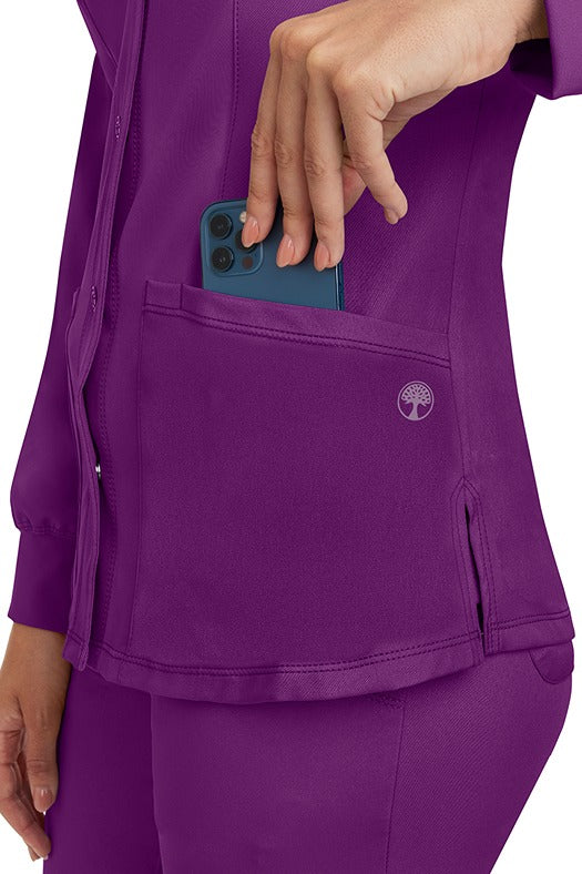 A young lady wearing an HH-Works Women's Megan Snap Front Scrub Jacket in Eggplant featuring two front patch pockets.