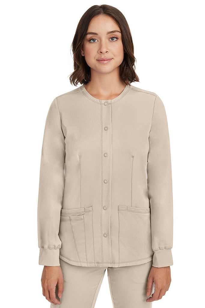 A young female nurse wearing a Women's Megan Snap Front Scrub Jacket from HH Works in Khaki featuring a round neckline & long sleeves.