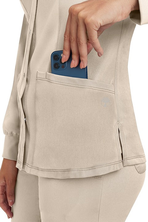 A young lady wearing an HH-Works Women's Megan Snap Front Scrub Jacket in Khaki featuring two front patch pockets.
