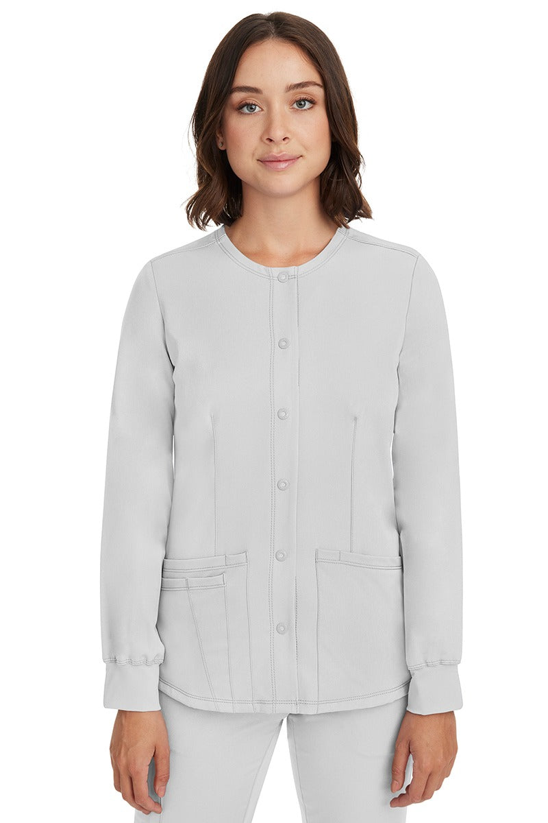 A young female nurse wearing a Women's Megan Snap Front Scrub Jacket from HH Works in White featuring a round neckline & long sleeves.