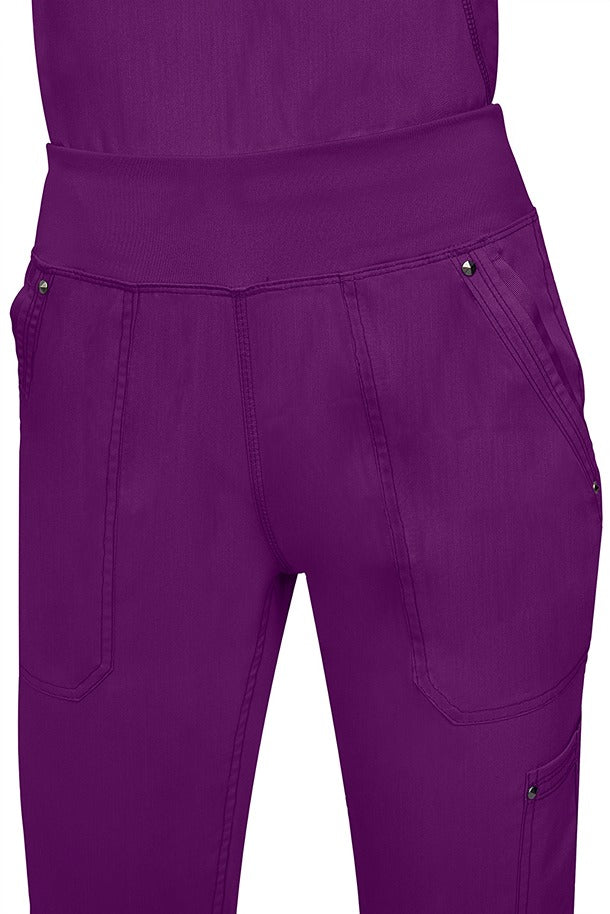 A young female healthcare professional wearing a Purple Label Women's Tori Yoga Waistband Scrub Pant in Eggplant featuring a unique fabric that resists wrinkles, shrinking, & fading better than traditional cotton scrubs.