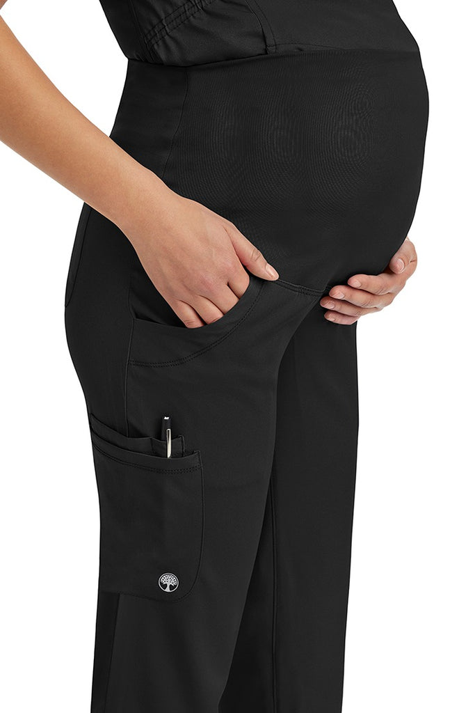 A young woman wearing an HH-Works Women's Rose Maternity Cargo Scrub Pant in Black featuring two side pockets and a double sided pocket for all of your on the hob storage needs.