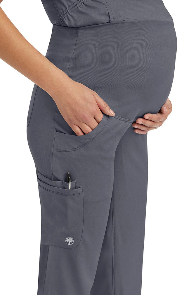 A young woman wearing an HH-Works Women's Rose Maternity Cargo Scrub Pant in Pewter featuring two side pockets and a double sided pocket for all of your on the hob storage needs.