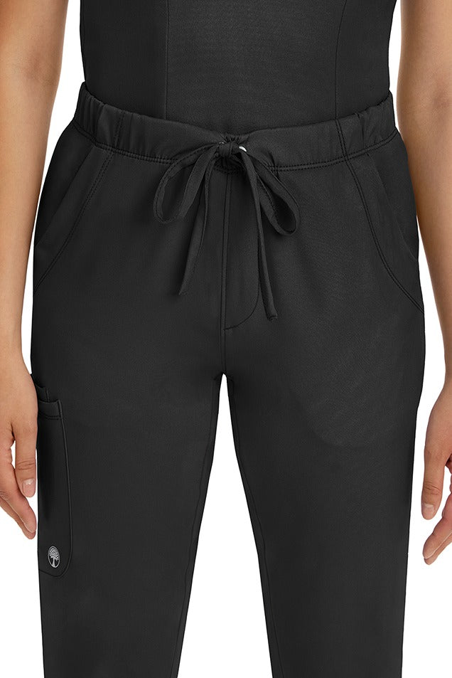 A female LPN wearing a pair of HH-Works Women's Rebecca Multi-Pocket Drawstring Pants in Black featuring 2 front slip pockets with a cargo pocket at the wearer's right leg. 