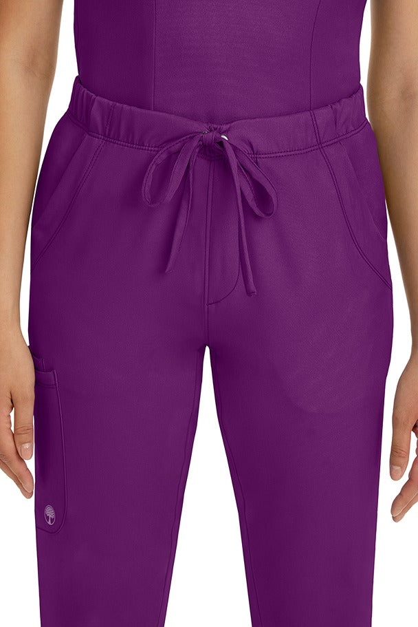 A female LPN wearing a pair of HH-Works Women's Rebecca Multi-Pocket Drawstring Pants in Eggplant featuring 2 front slip pockets with a cargo pocket at the wearer's right leg.