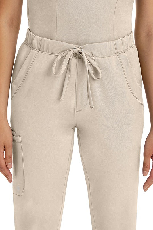 A female LPN wearing a pair of HH-Works Women's Rebecca Multi-Pocket Drawstring Pants in Khaki featuring 2 front slip pockets with a cargo pocket at the wearer's right leg.