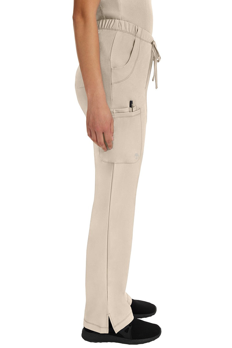 A young female nurse wearing an HH-Works Women's Rebecca Multi-Pocket Drawstring Pant in Khaki featuring side vents at the hem to provide additional range of motion throughout the day.