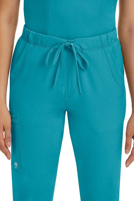A female LPN wearing a pair of HH-Works Women's Rebecca Multi-Pocket Drawstring Pants in Teal featuring 2 front slip pockets with a cargo pocket at the wearer's right leg.